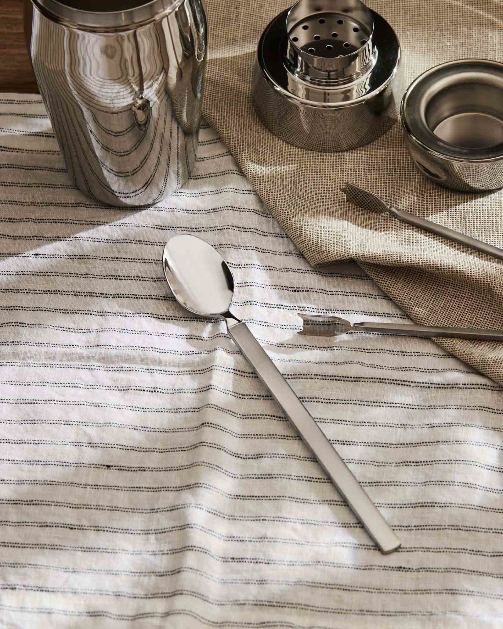 Stainless steel spoon by Alessi with a polished bowl and matte handle, on a striped white linen cloth.