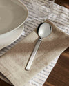 Silver-toned serving spoon by Alessi with a polished bowl and brushed handle, on a textured beige napkin.