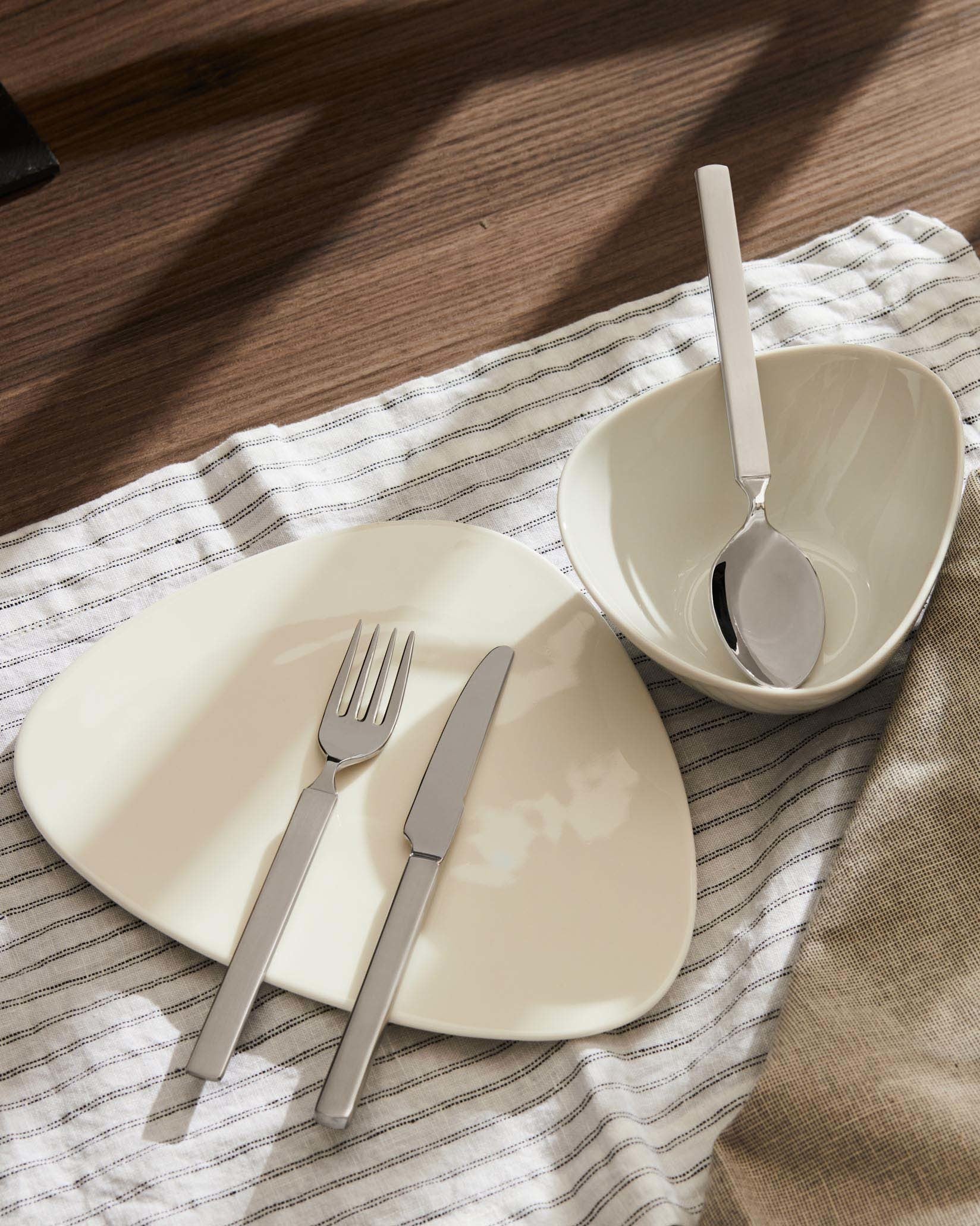 Dry stainless steel cutlery set by Alessi, on a cream irregular-shaped plate with striped and beige linen, on a wood table.