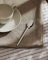 Polished silver spoon by Alessi with a sleek, faceted handle, shown on a woven placemat with a white cup.