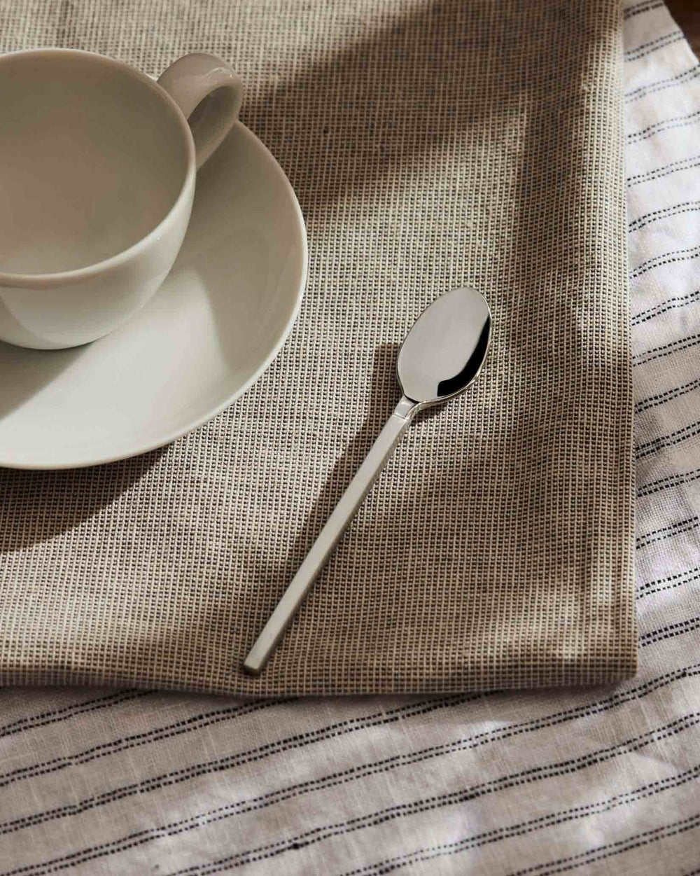 Silver metal spoon by Alessi resting on a beige woven placemat with a white striped cloth and ceramic cup.