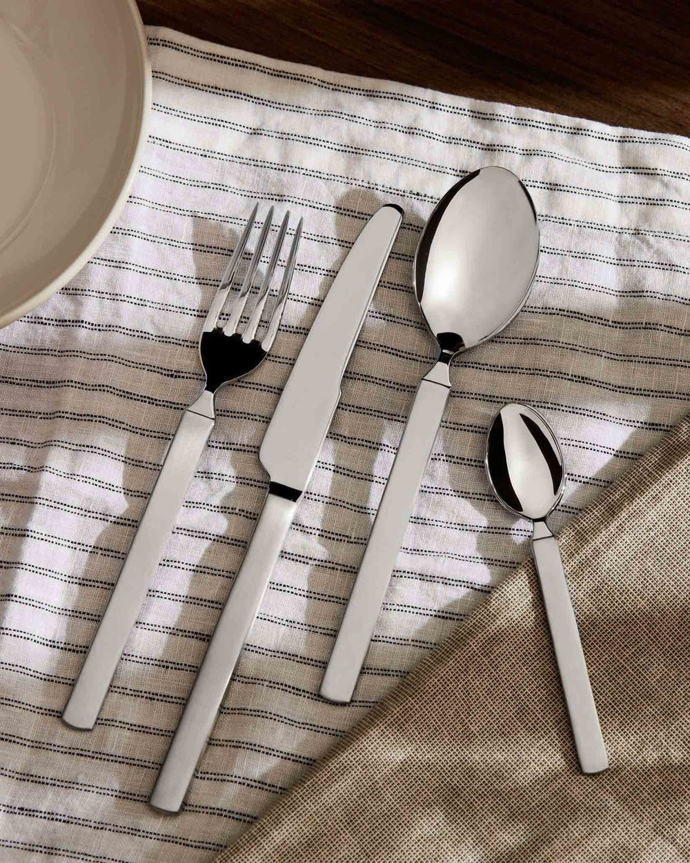 Sleek stainless steel cutlery set by Alessi, laid on a striped white napkin with a plate.