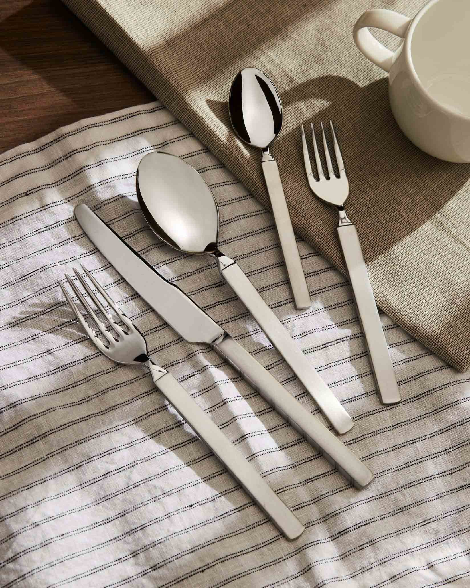 Stainless steel cutlery set with white handles by Alessi, including knife, fork, and two spoons on a striped napkin.