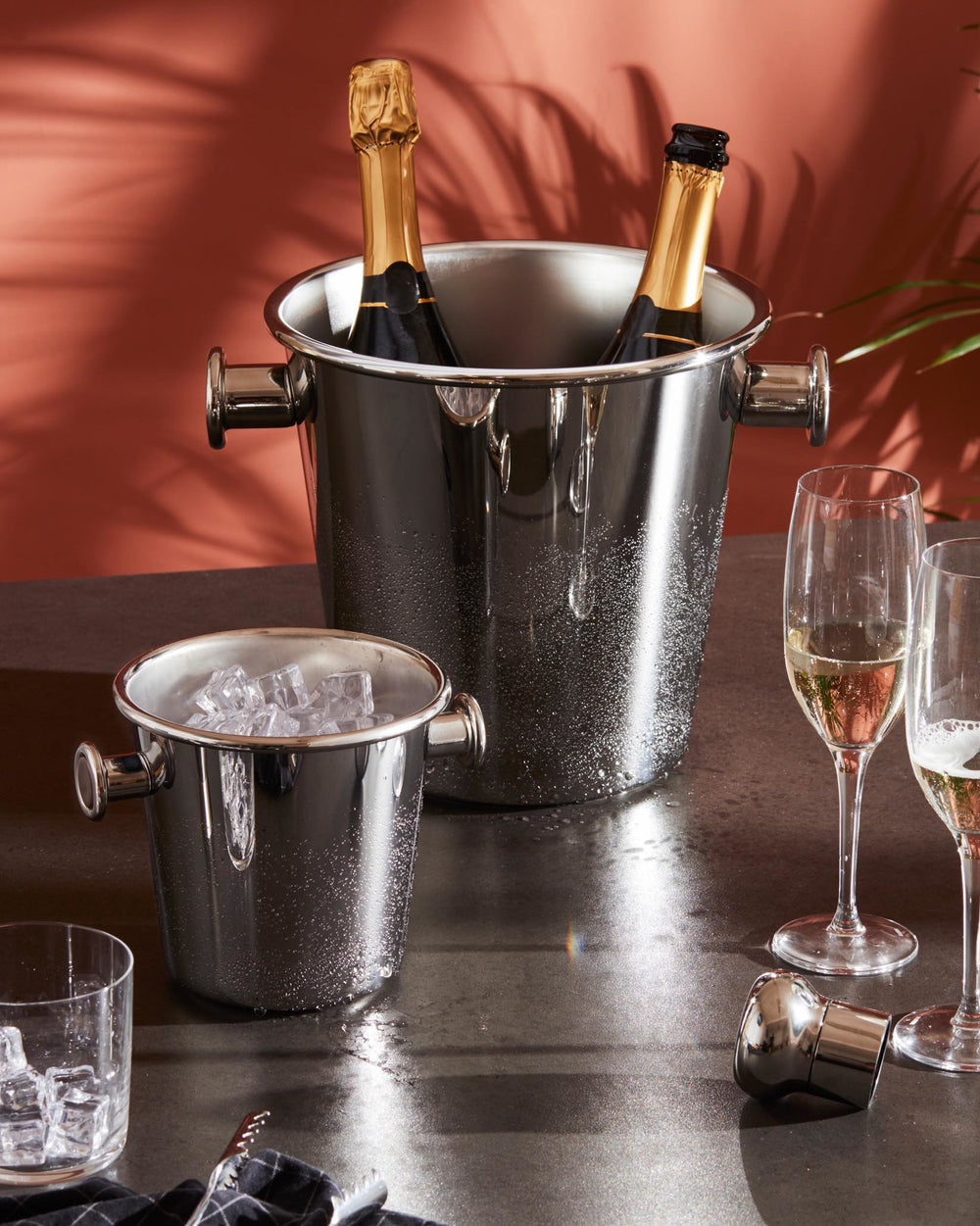 Silver metallic bottle stopper by Alessi, lying on a dark surface next to champagne flutes.