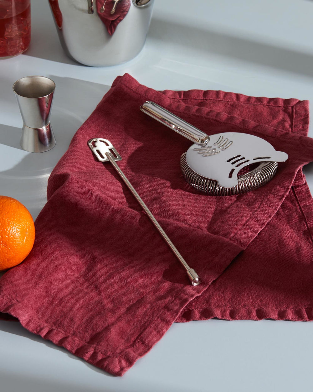 Silver metal cocktail shaker by Alessi, with a strainer, stirring spoon, and jigger on a red linen napkin.