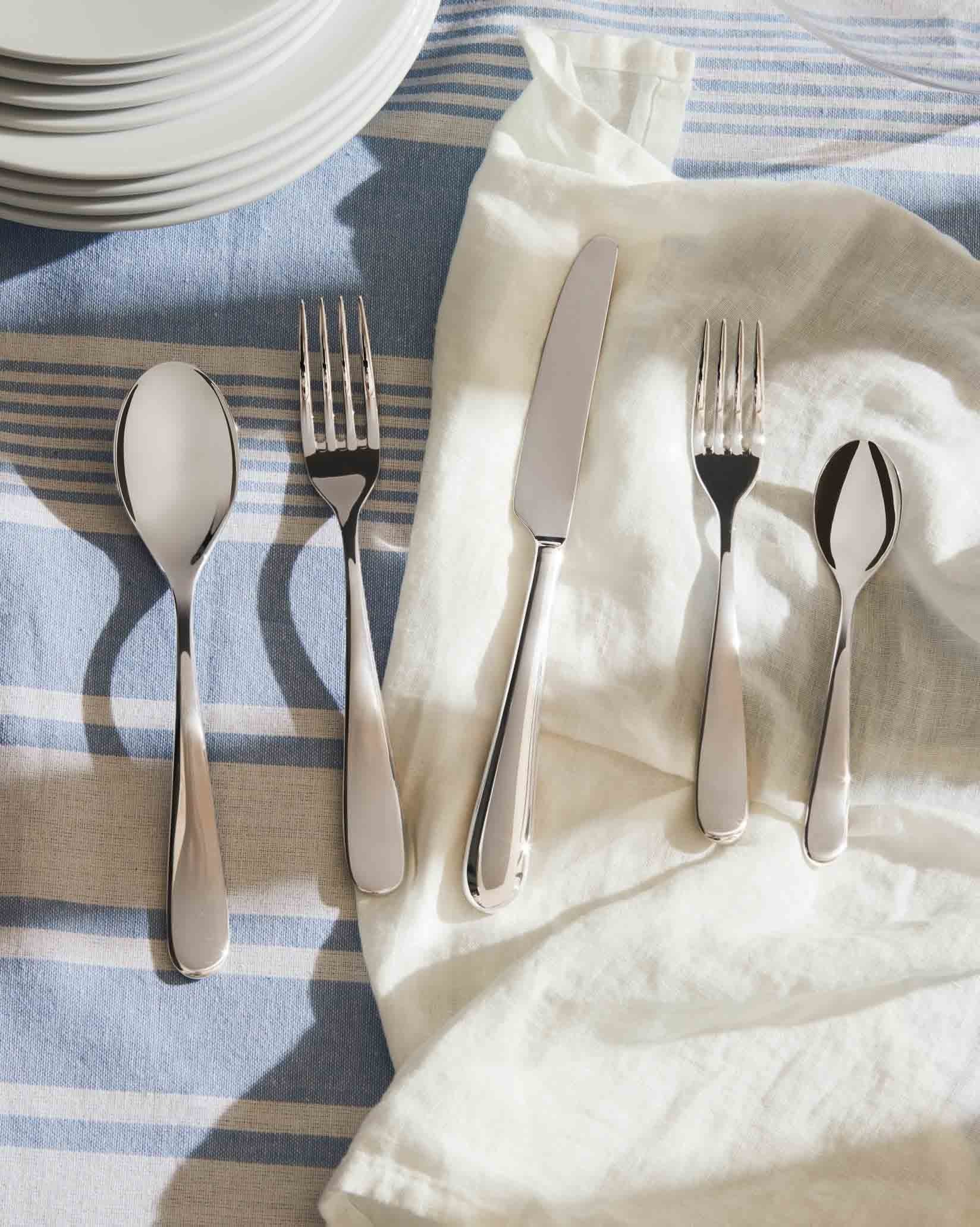 Five-piece stainless steel flatware set by Alessi on a blue and white striped tablecloth with a white napkin.