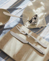 Polished stainless steel serving utensils by Alessi, including a fork, knife, and ladle, arranged on a marble board with a striped linen cloth.