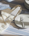 Silver-toned cake server by Alessi, with a wide flat head, resting on a white napkin over a blue striped tablecloth.