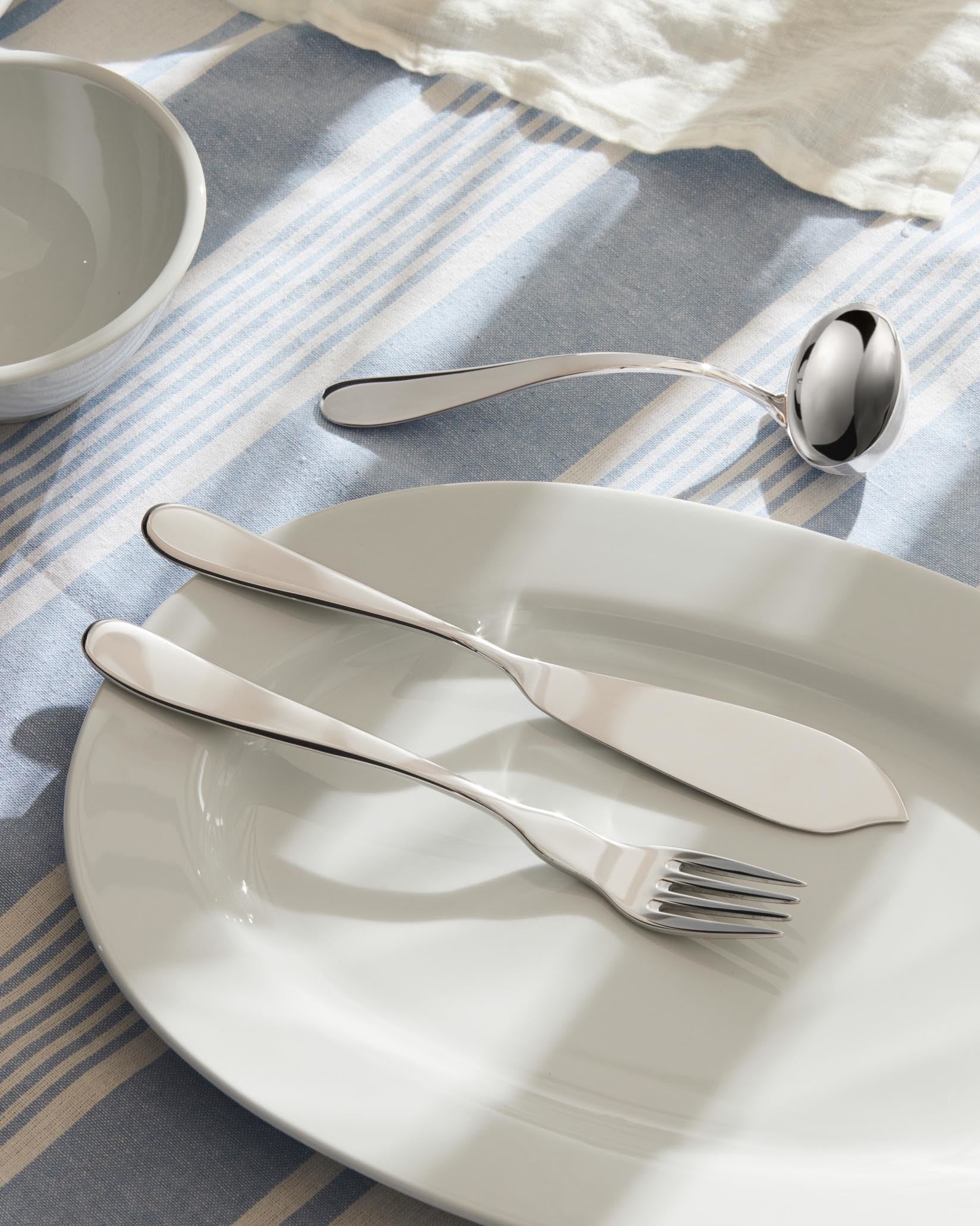 Sleek silver-toned fork, knife, and ladle by Alessi, on a white oval plate and a striped tablecloth.