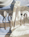 Five polished silver-colored cutlery pieces by Alessi, with sleek design, arranged on a striped tablecloth and white napkin.
