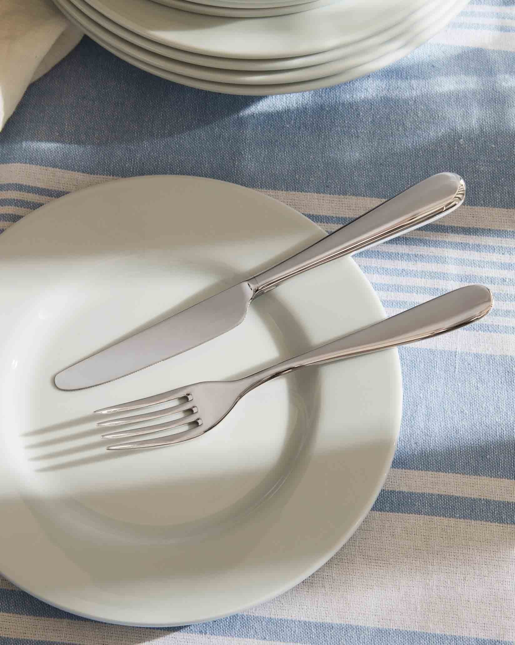 Silver fork and knife by Alessi resting on a white plate on a blue and white striped tablecloth.