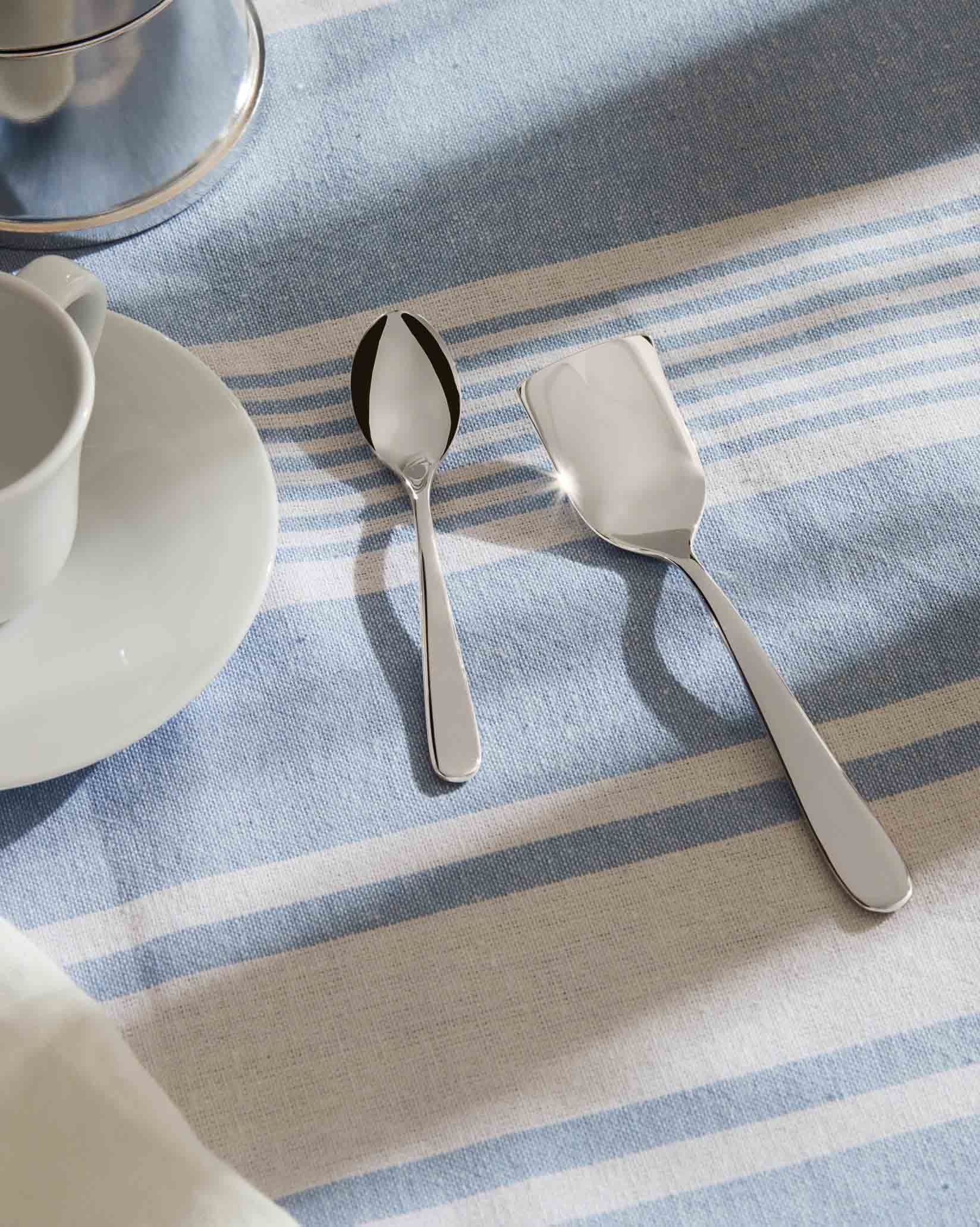 Sleek silver-colored cutlery by Alessi, featuring a spoon and a rectangular server on a striped tablecloth.