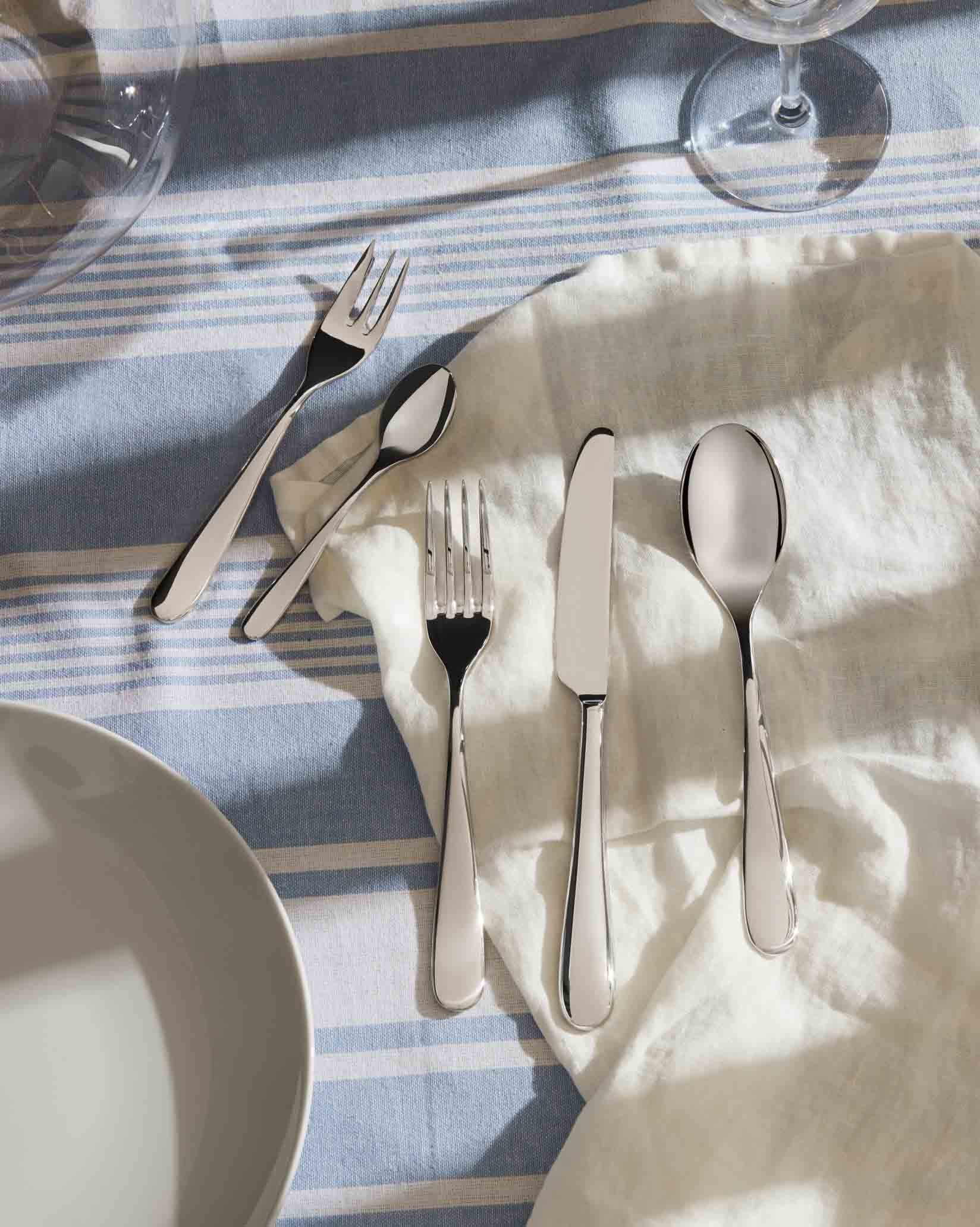 Polished silver cutlery set by Alessi, laid on a blue and white striped tablecloth with a white linen napkin.