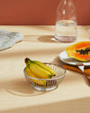 Silver metal wire fruit basket by Alessi, holding yellow bananas on a light wood table.
