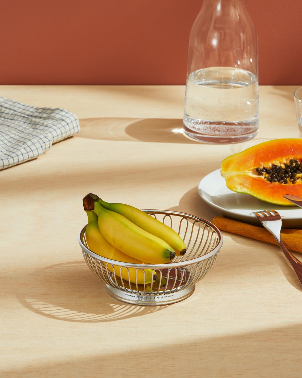 Silver metal wire fruit basket by Alessi, holding yellow bananas on a light wood table.