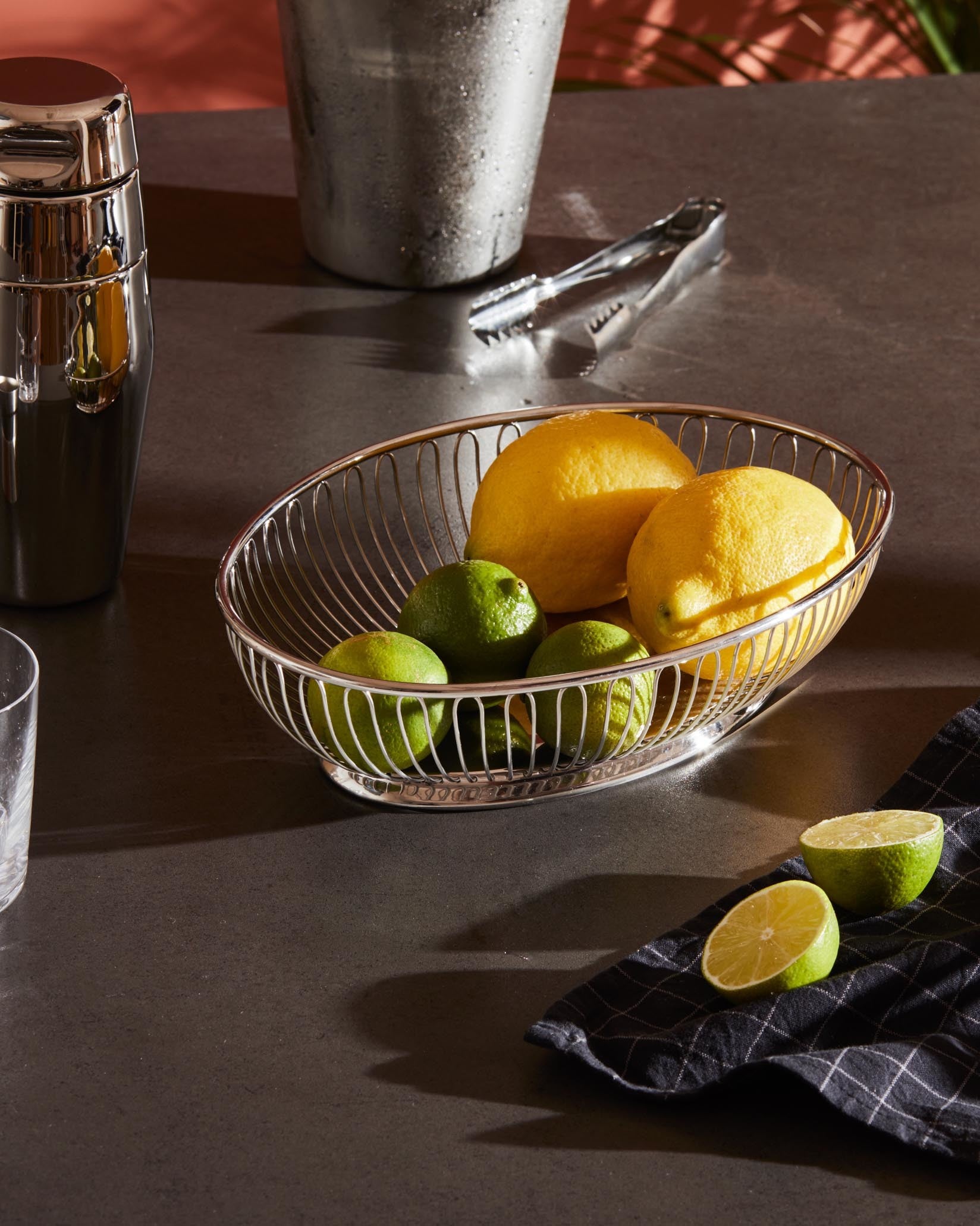 Silver metal oval wire fruit bowl by Alessi, holding lemons and limes on a dark counter.