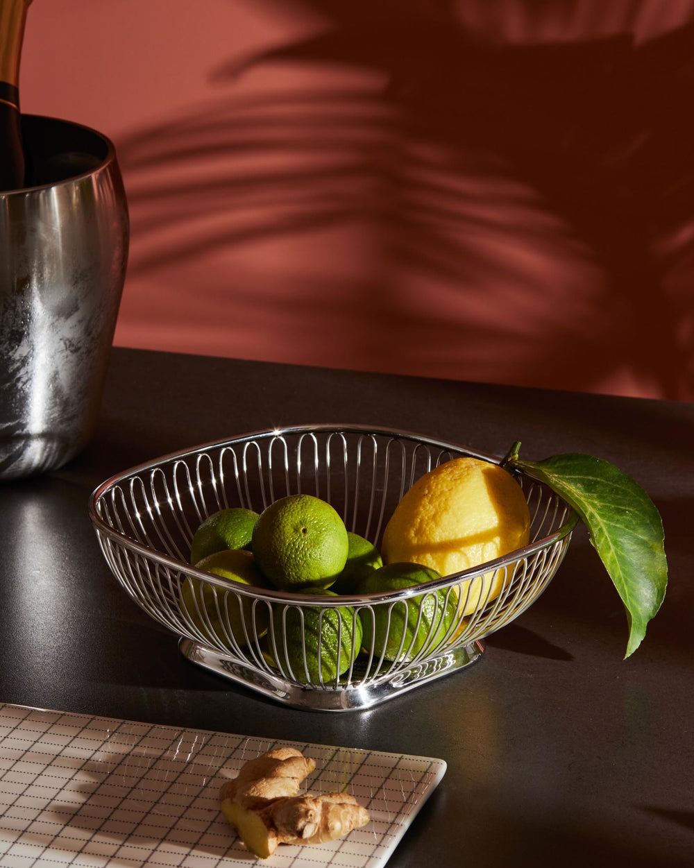 Silver wire fruit basket by Alessi, filled with limes and a lemon on a dark counter with ginger.
