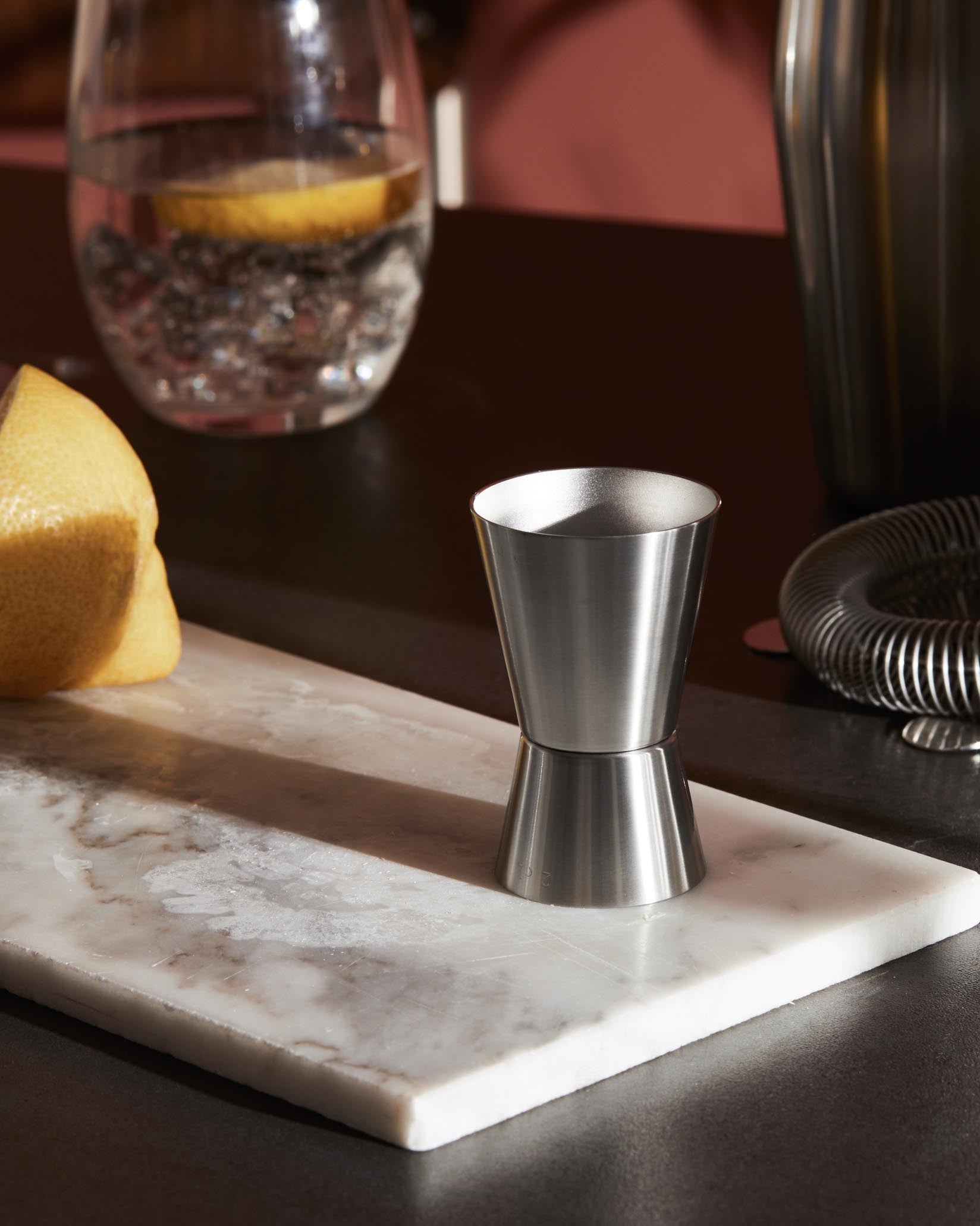 Brushed stainless steel double jigger by Alessi, standing on a white marble slab next to a lemon and a glass of water.