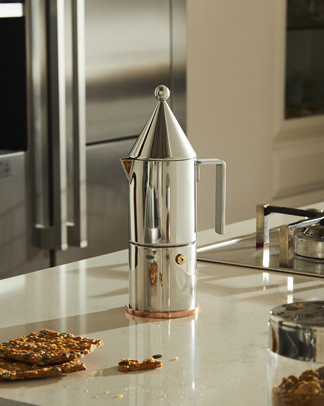 Silver conical espresso maker by Alessi, with a pointed top, finial, and copper base on a kitchen counter.