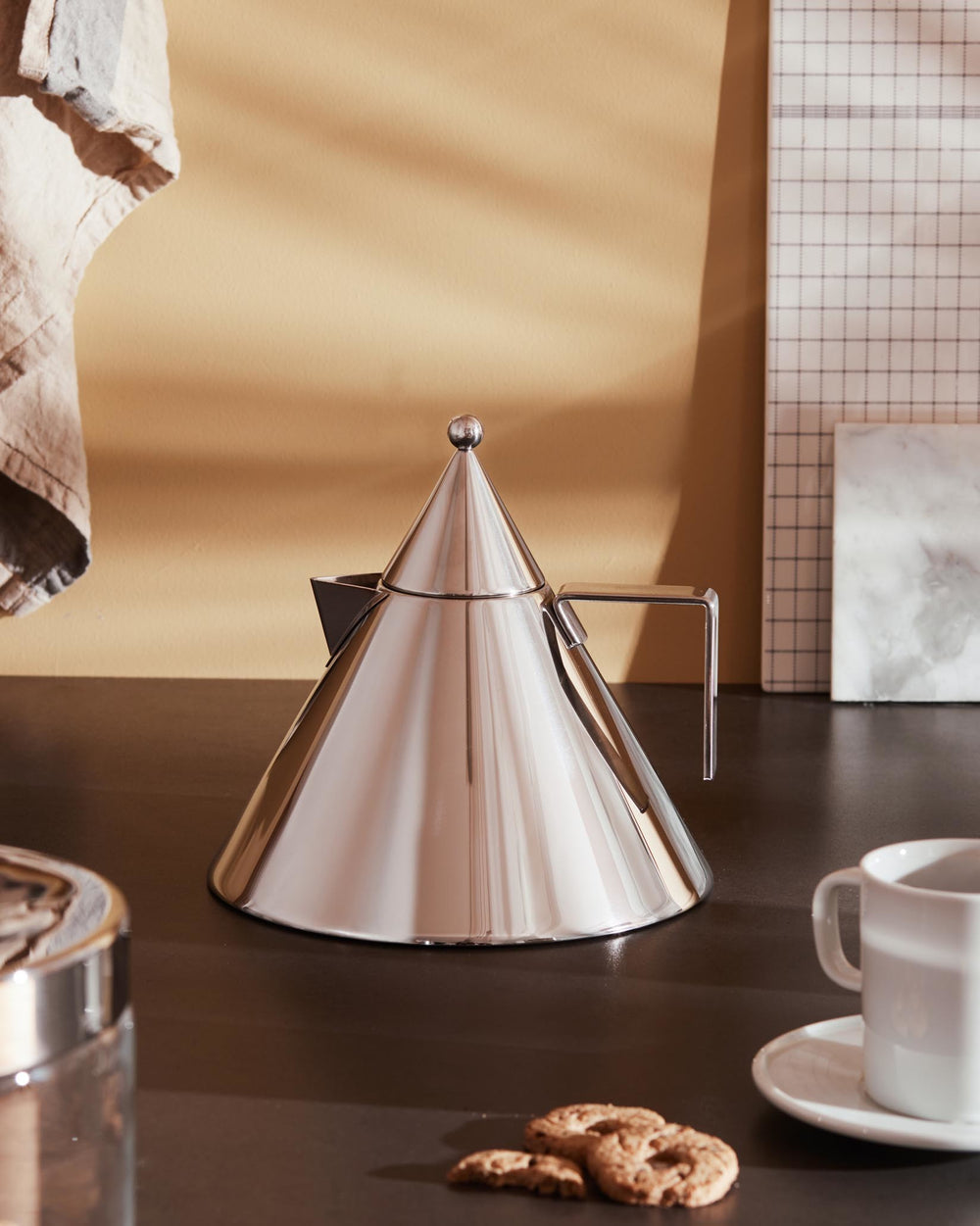 Polished stainless steel conical coffee maker by Alessi, with a spherical knob and U-shaped handle, on dark counter.