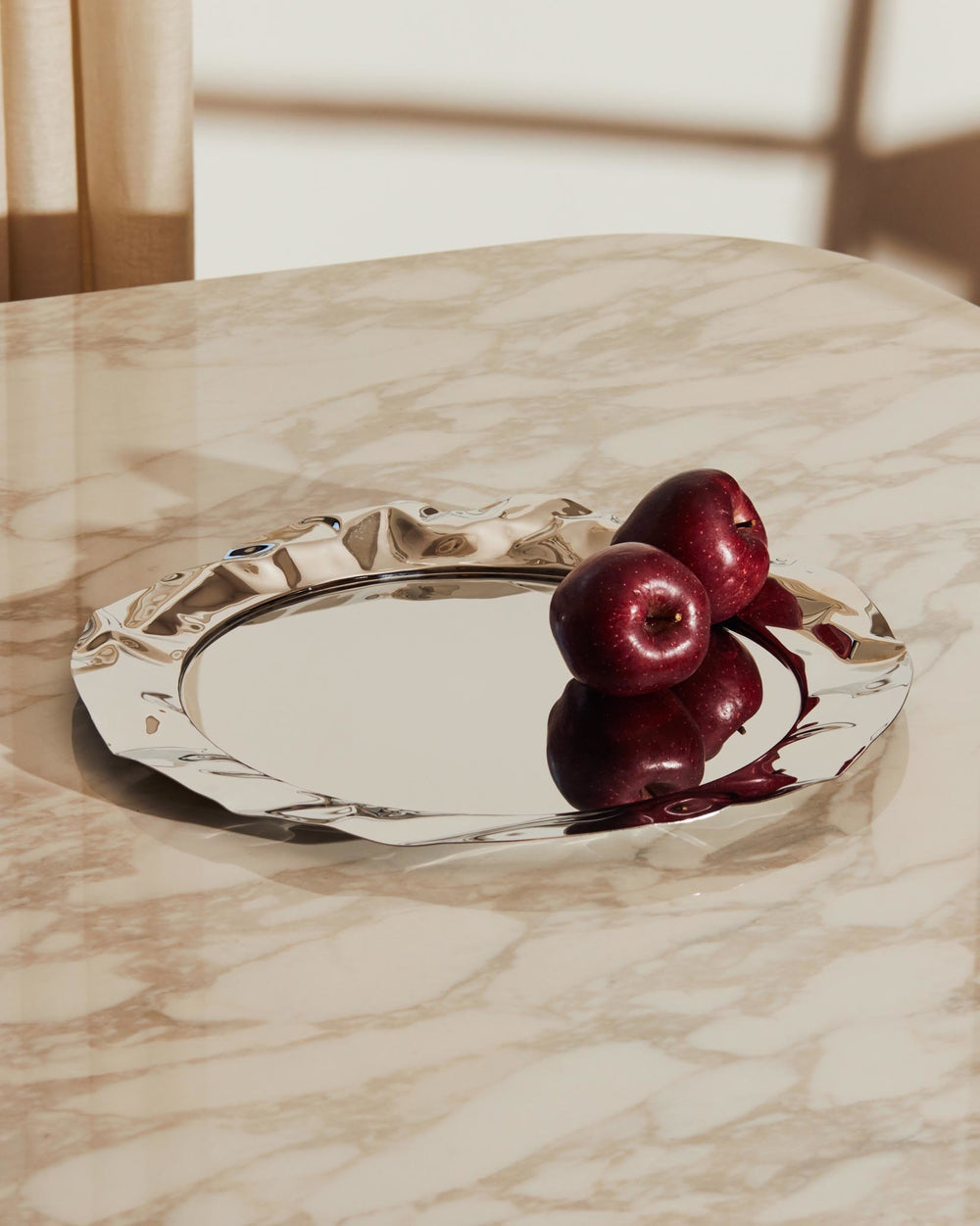 Silver polished tray with a wavy edge by Alessi, reflecting red apples on a marble surface.