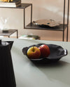Black matte fruit bowl by Alessi with wavy edges, holding two apples on a light grey counter.