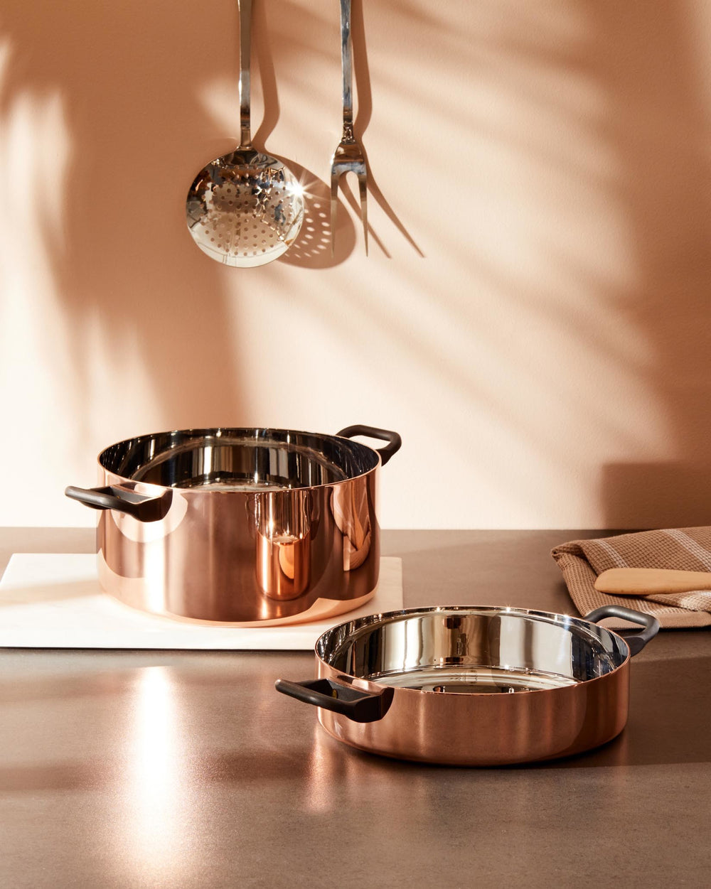 Copper cookware set with black handles by Alessi, featuring a large pot and shallow pan on a kitchen counter.