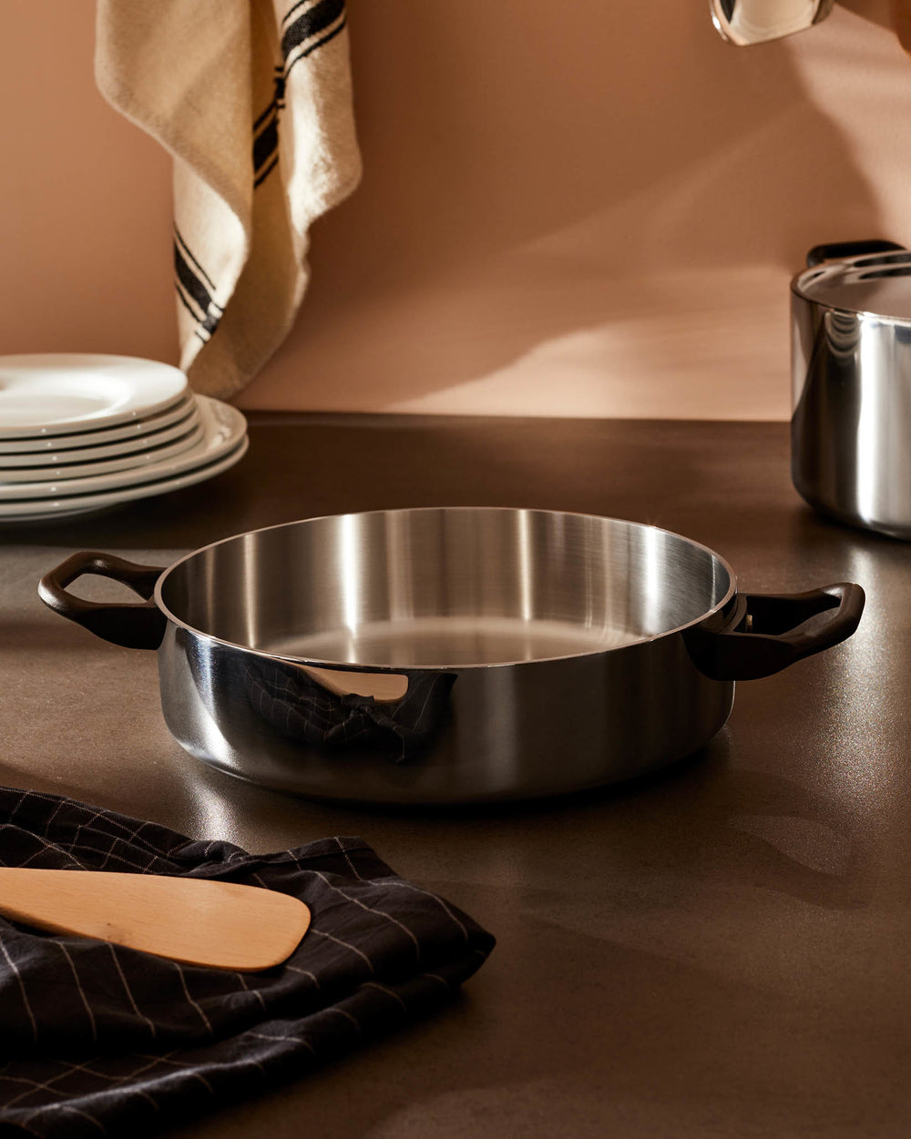 Stainless steel sauté pan La Cintura di Orione by Alessi, with black handles, a wooden spatula, and stacked white plates.