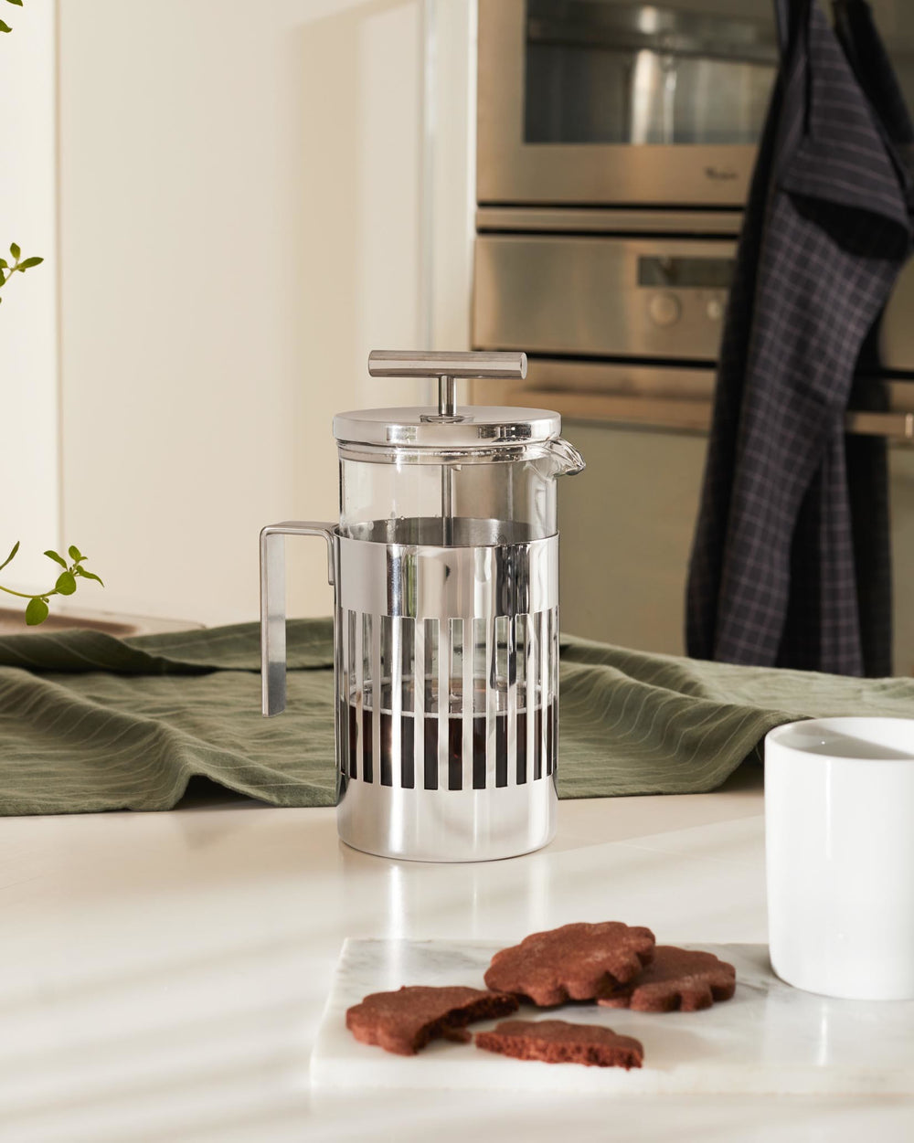 Stainless steel and glass French press with vertical bars by Alessi, filled with dark coffee, on a kitchen counter.