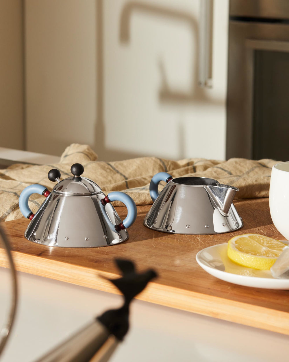 Polished stainless steel conical tea kettle by Alessi with light blue handles and black knobs, alongside a matching creamer.
