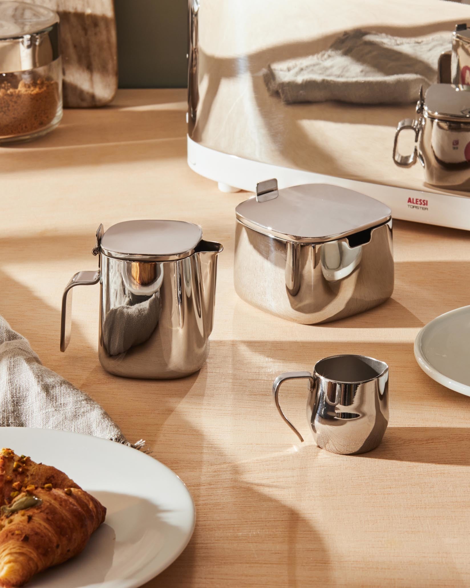 Silver stainless steel sugar bowl by Alessi, with matching creamer and small milk jug on a wooden table with a croissant.