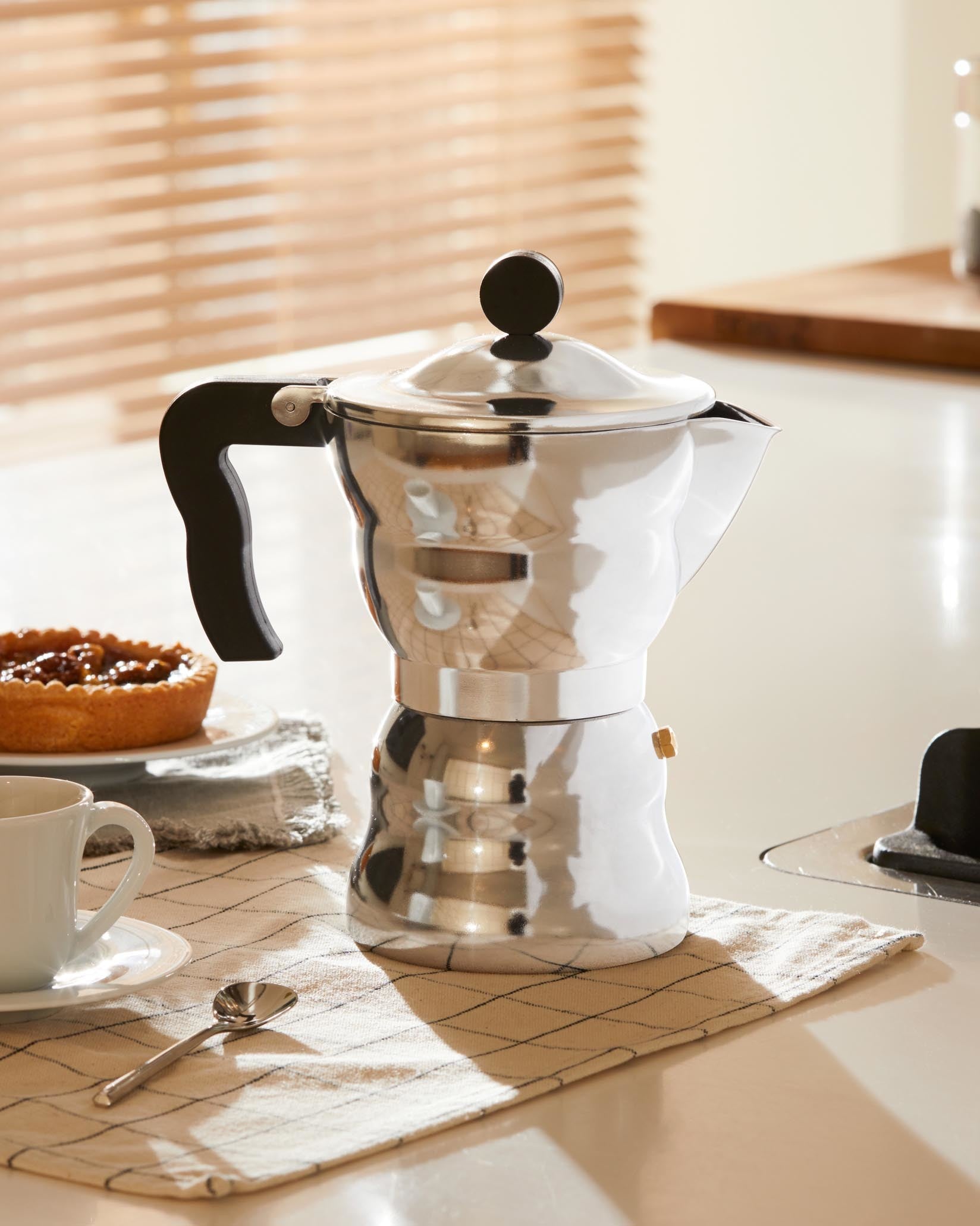 Polished silver Moka pot by Alessi with a black handle and knob, on a kitchen counter with a pastry and cup.
