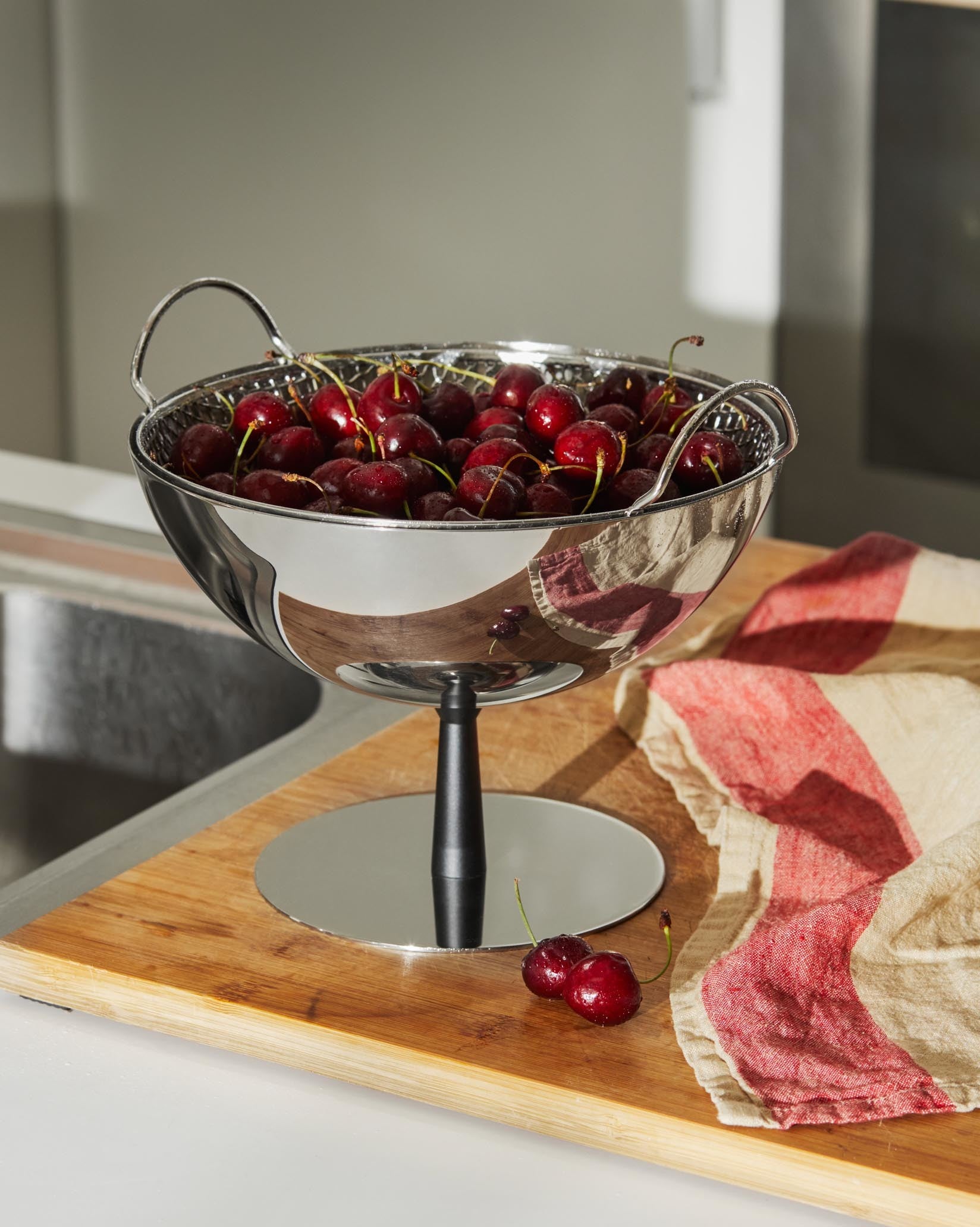 Silver fruit bowl with perforated insert and black stand by Alessi, filled with cherries on a wooden board and striped towel.