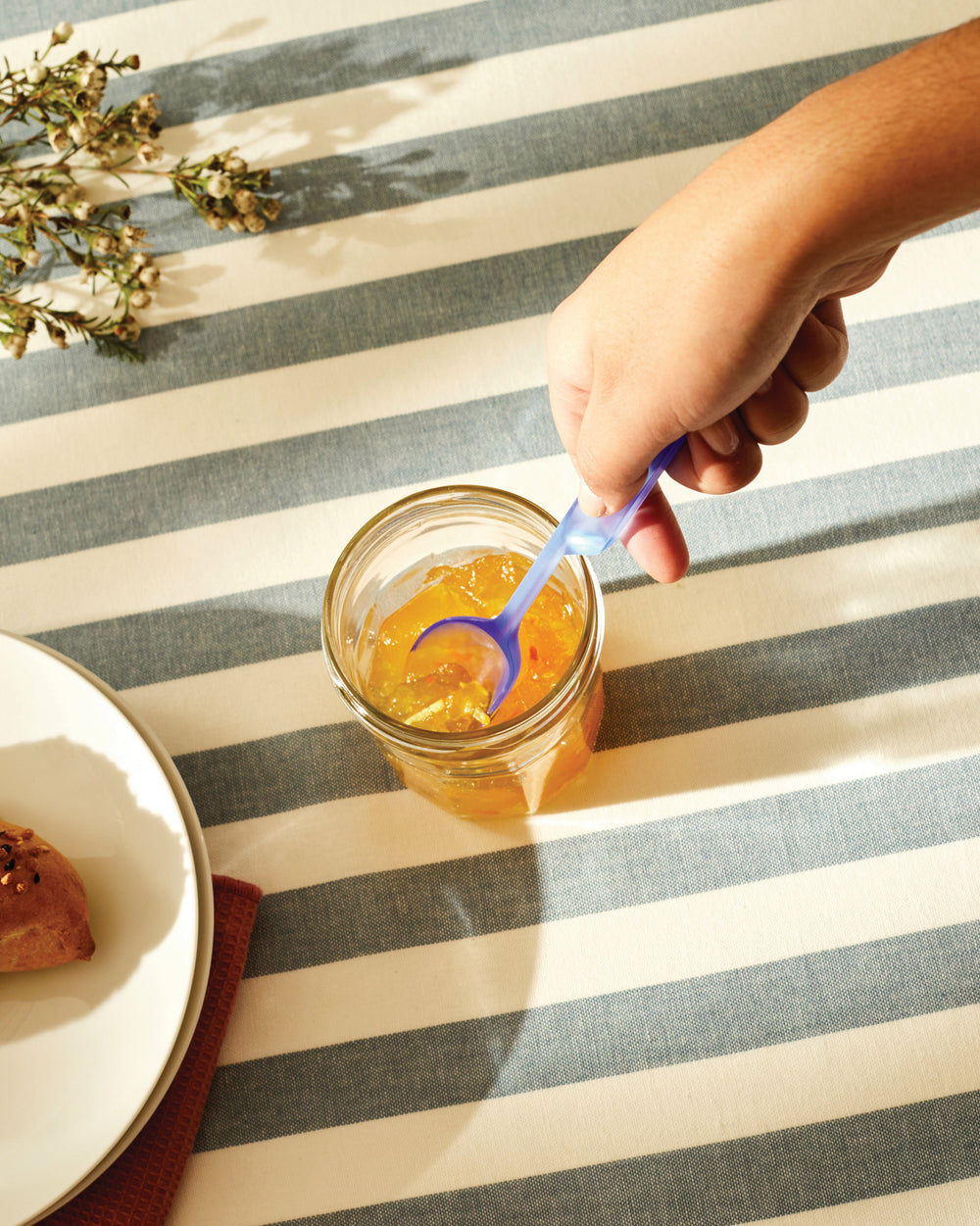 Blue plastic spoon by Alessi stirring orange marmalade in a glass jar on a striped tablecloth.