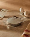 Silver metallic geometric egg cups by Alessi, holding white eggs on a speckled counter with a shiny pan, spatula, and striped towel.