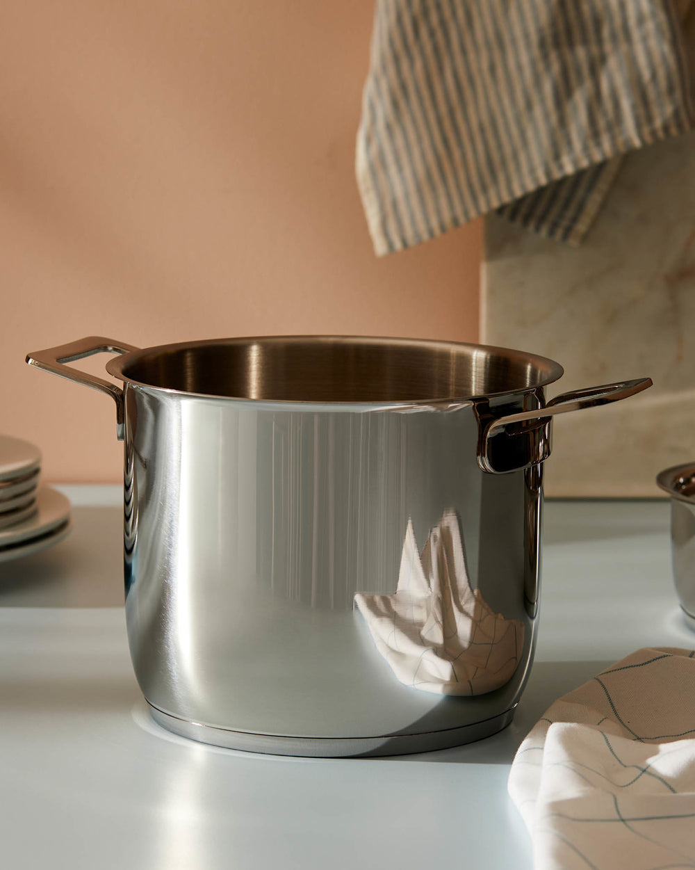 Polished stainless steel pot by Alessi with two handles, reflecting a striped towel on a light counter.