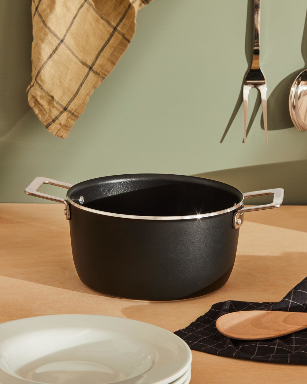 Black casserole pot by Alessi with silver handles and rim, on a wooden surface with kitchen tools.
