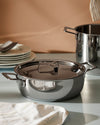 Stainless steel pot with lid by Alessi, featuring sleek handles and reflections on a light surface with kitchen items.