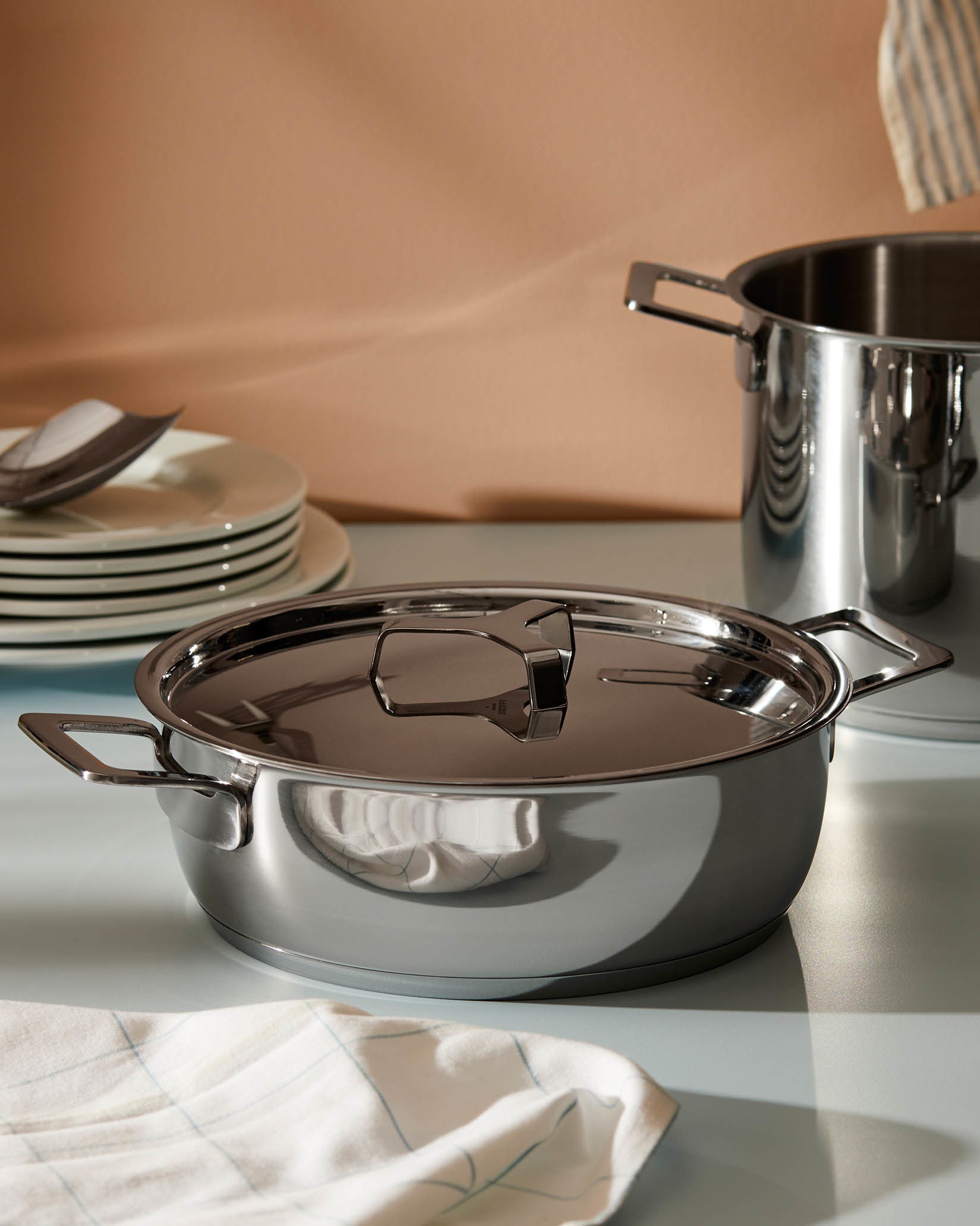 Stainless steel pot with lid by Alessi, featuring sleek handles and reflections on a light surface with kitchen items.
