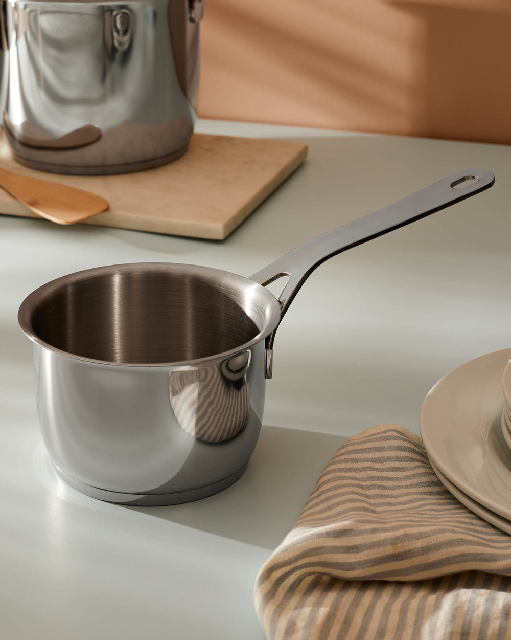 Silver stainless steel saucepan by Alessi with a long handle, resting on a light grey countertop with kitchen items.