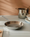 Stainless steel frying pan and stockpot by Alessi on a light grey counter, next to stacked white plates.