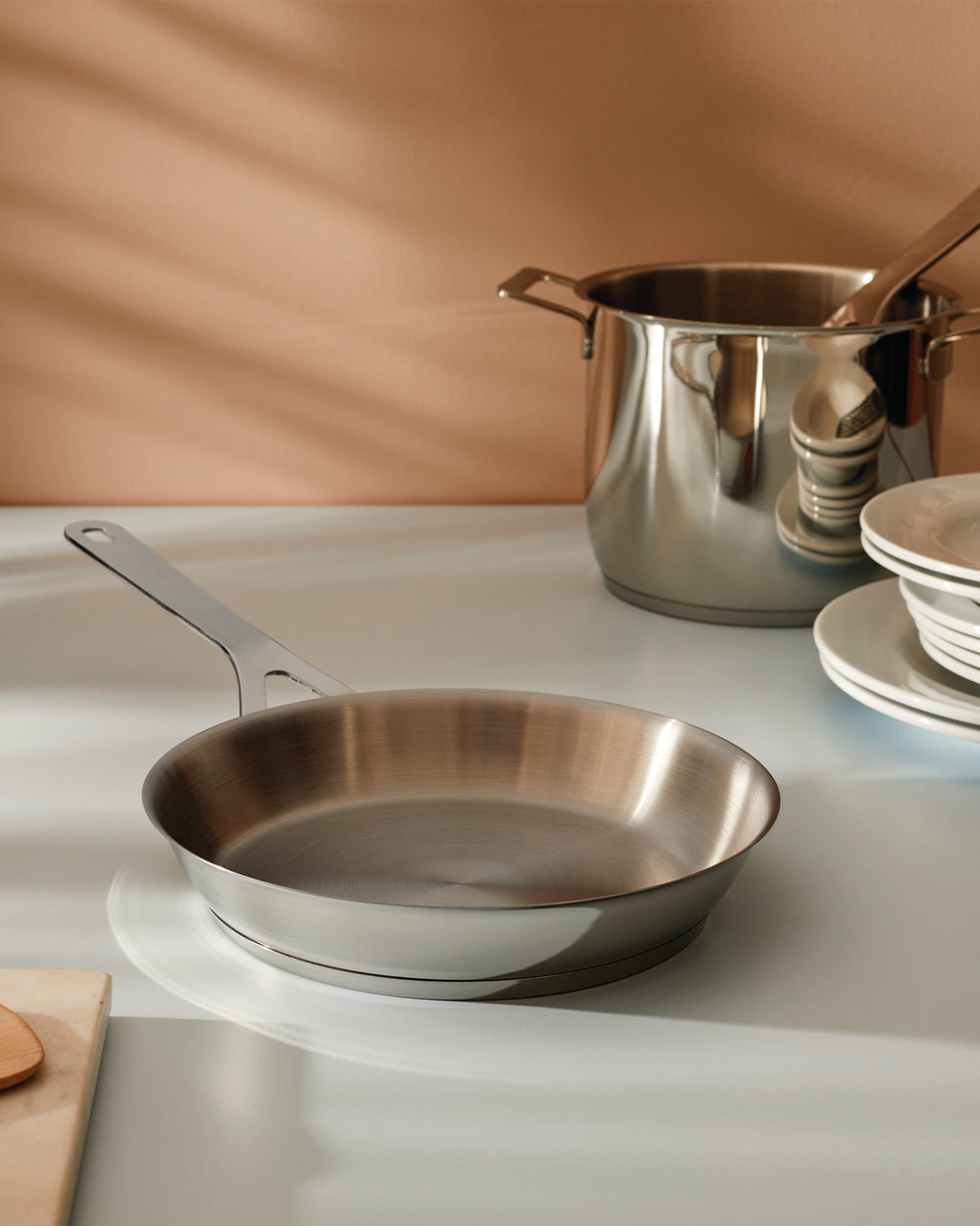 Stainless steel frying pan and stockpot by Alessi on a light grey counter, next to stacked white plates.