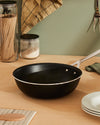 Black non-stick pan by Alessi with a shiny silver handle on a light wood kitchen counter.