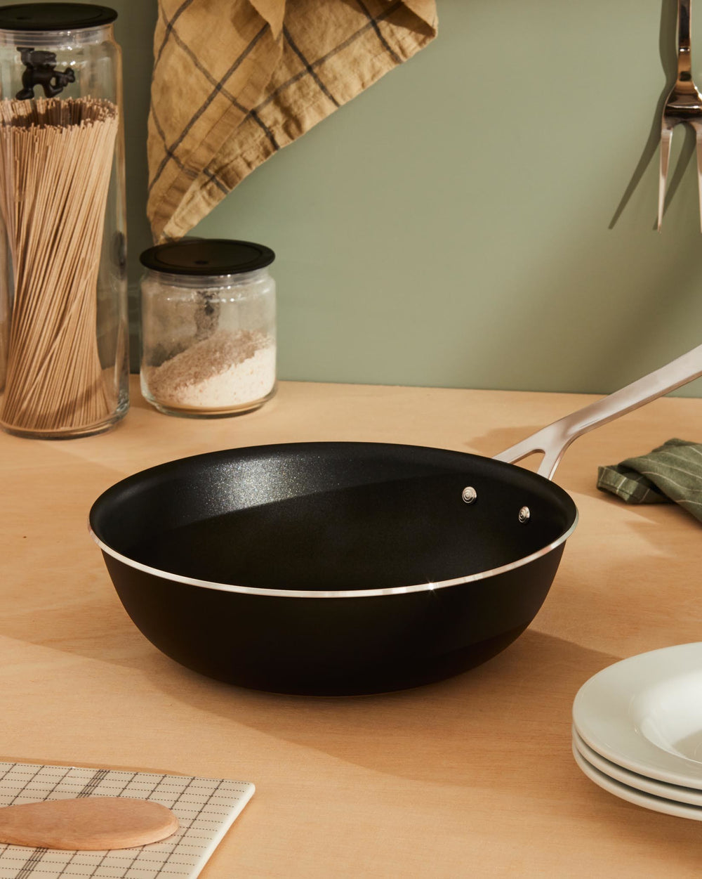 Black non-stick pan by Alessi with a shiny silver handle on a light wood kitchen counter.