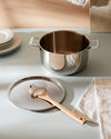 Stainless steel cooking pot by Alessi with handles, a matching lid holding a wooden spoon, stacked plates, and a checkered towel.