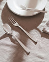 Sleek silver spoon and two forks by Alessi, styled on a light brown linen tablecloth with white plates.