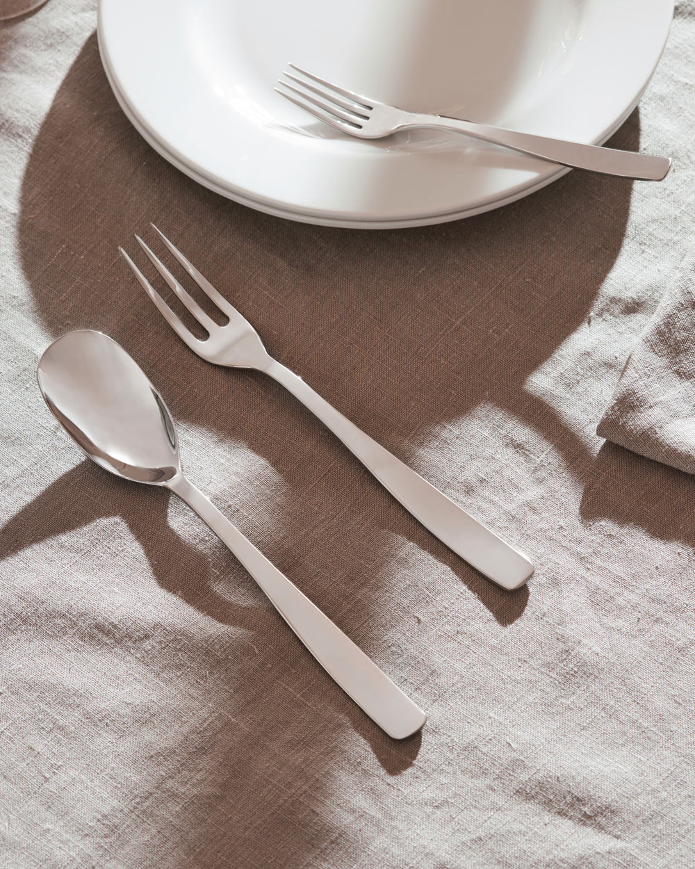 Sleek silver spoon and two forks by Alessi, styled on a light brown linen tablecloth with white plates.