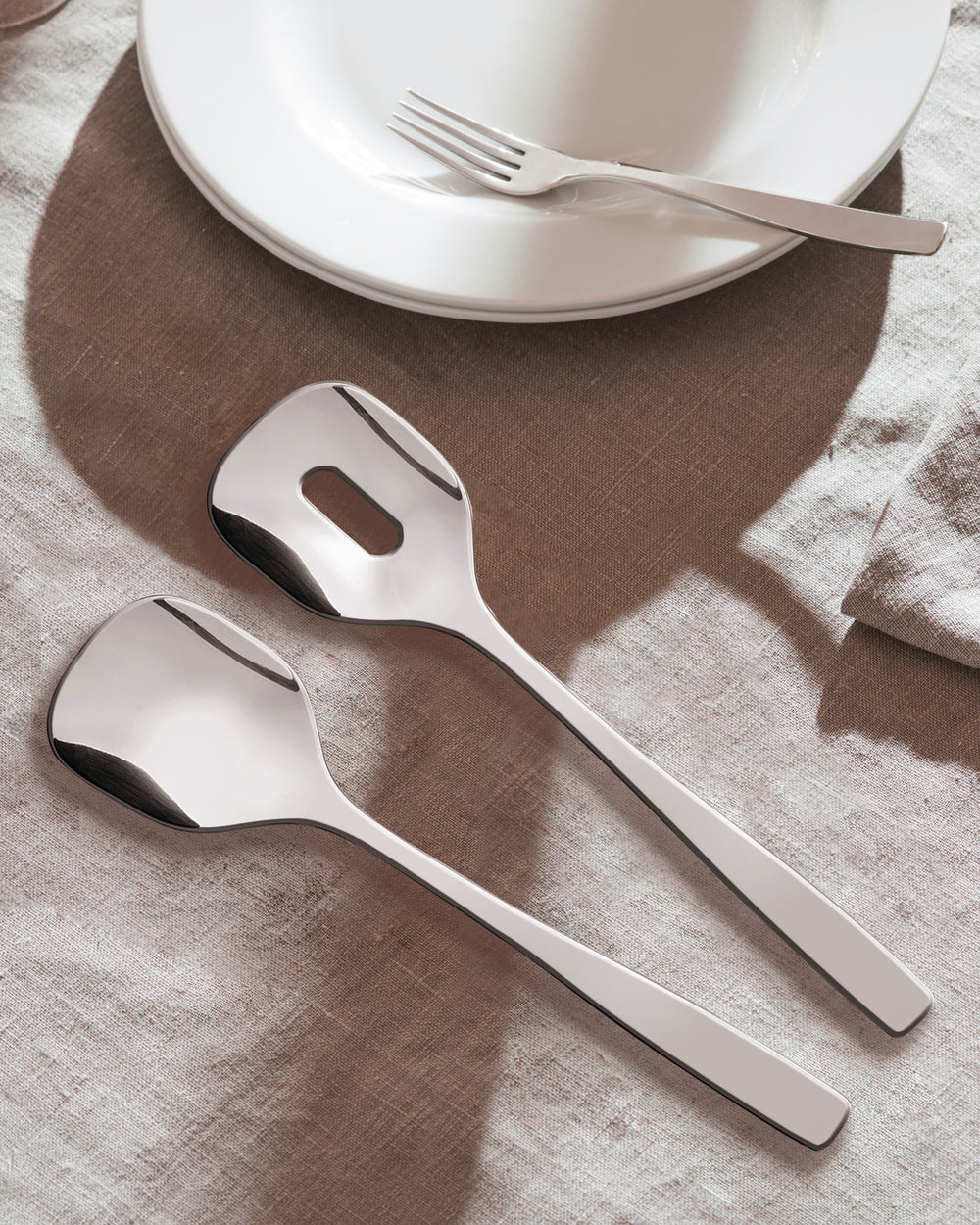 Shiny silver serving utensils by Alessi, one slotted, with a dinner fork on white plates.
