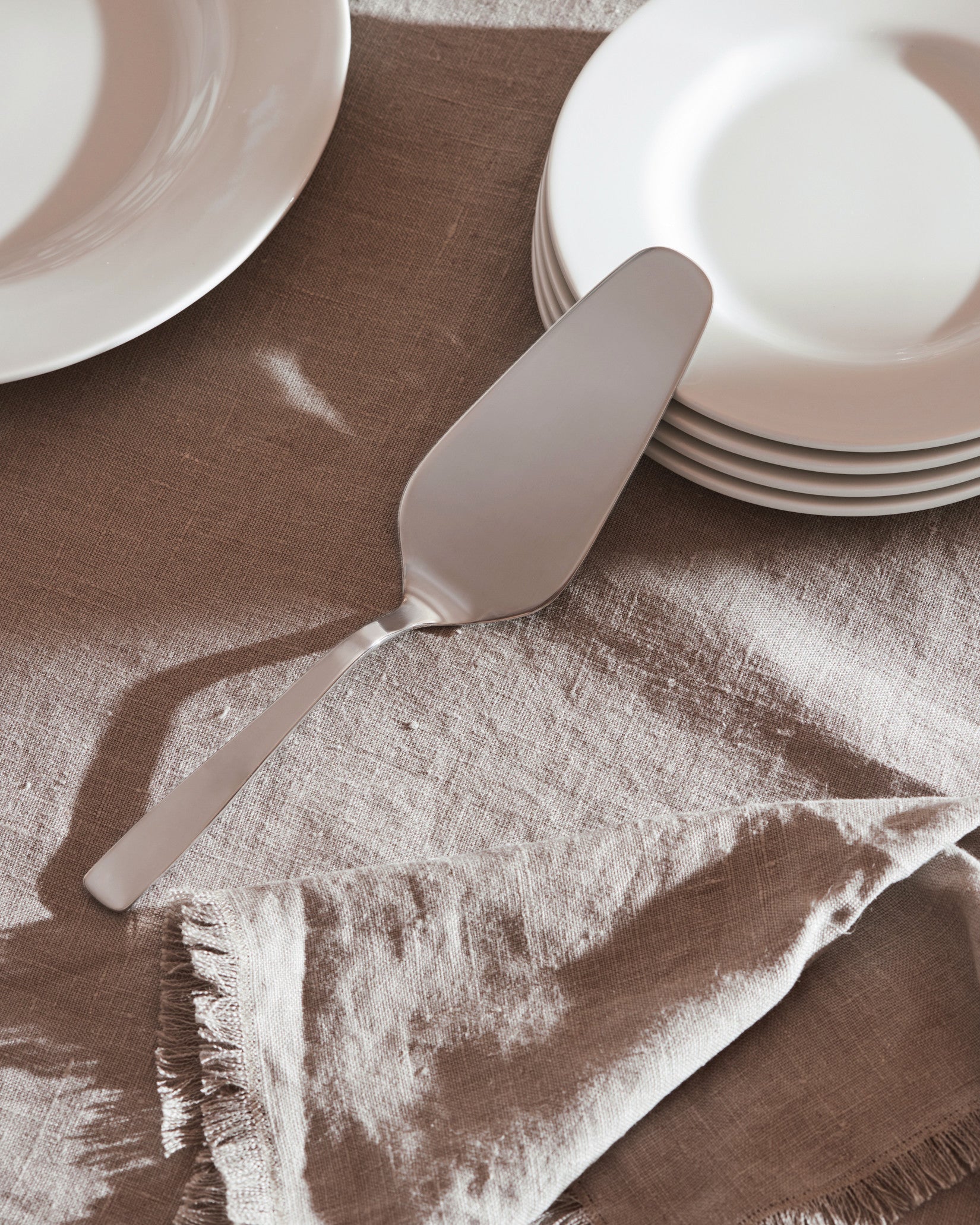 Silver-colored stainless steel cake server by Alessi, resting on a textured linen tablecloth next to white plates and a fringed napkin.
