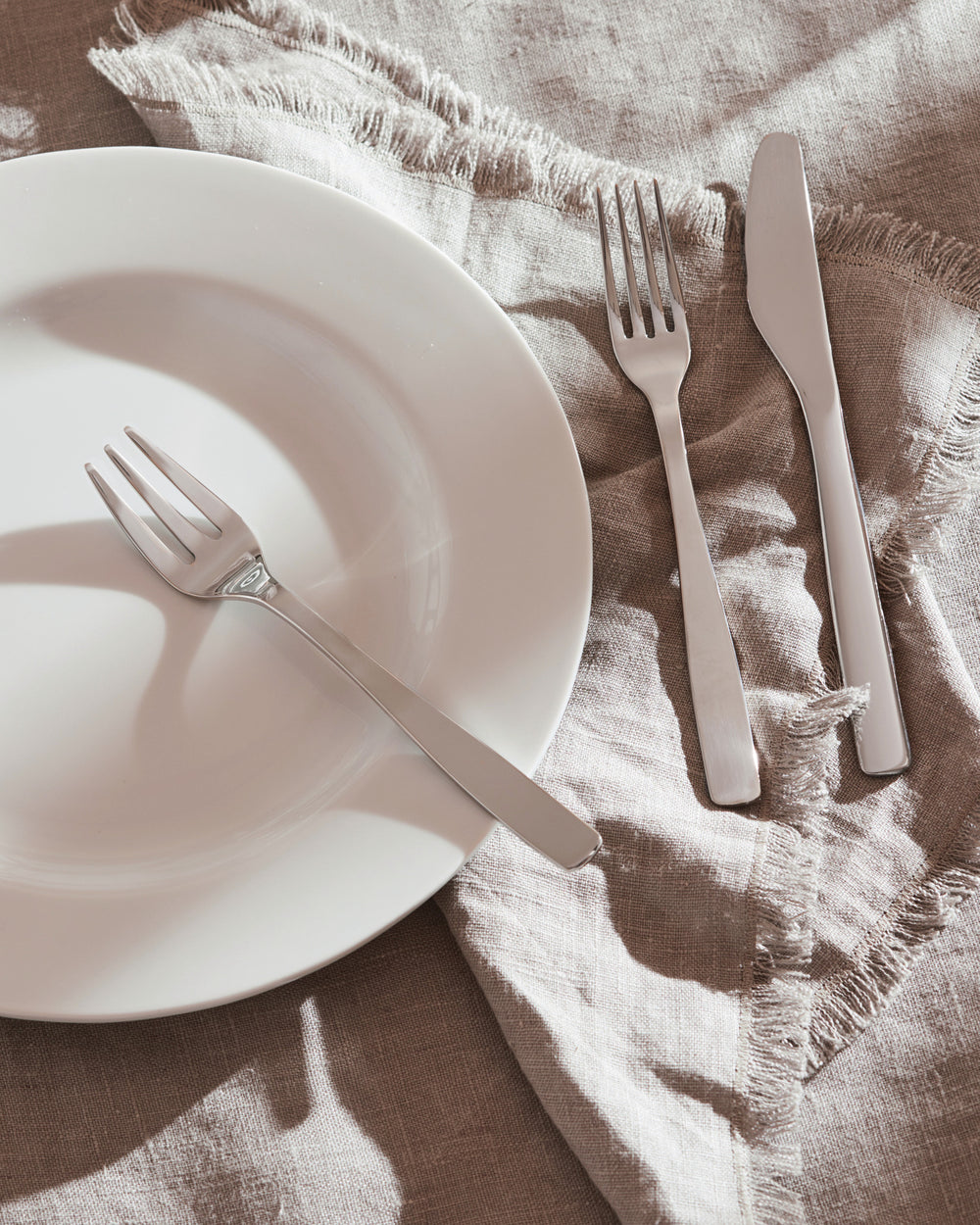 Silver metal KnifeForkSpoon set by Alessi, with a fork on a white plate and a knife and fork on a frayed linen napkin with sharp shadows.